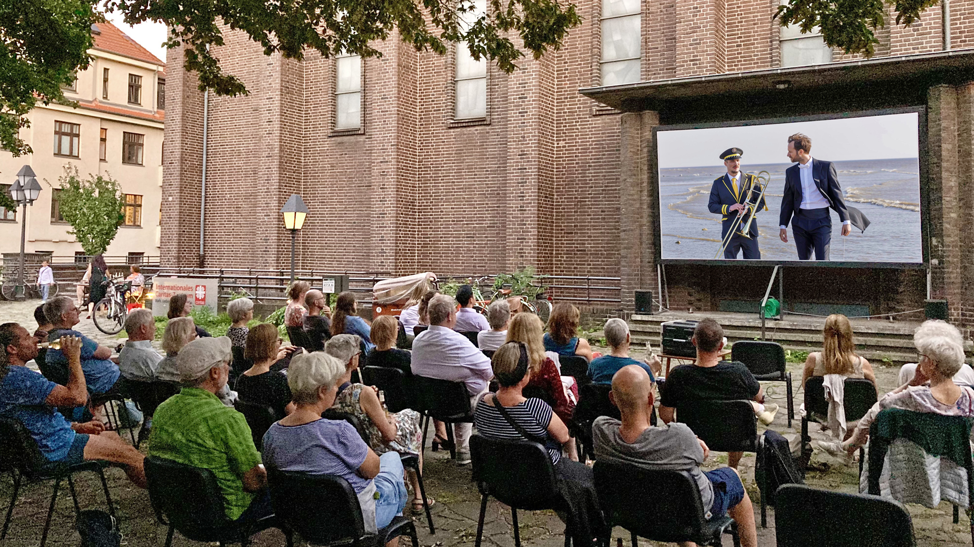 Eine Gruppe von Menschen sitzt auf Stühlen im Freien vor einer großen Leinwand und schaut einen Film. Die Vorführung findet in einem Hof neben einem Backsteingebäude statt. Auf der Leinwand sind zwei Männer im Anzug zu sehen, die am Strand entlanggehen; einer trägt zusätzlich eine Uniformmütze und ein Posauneninstrument.