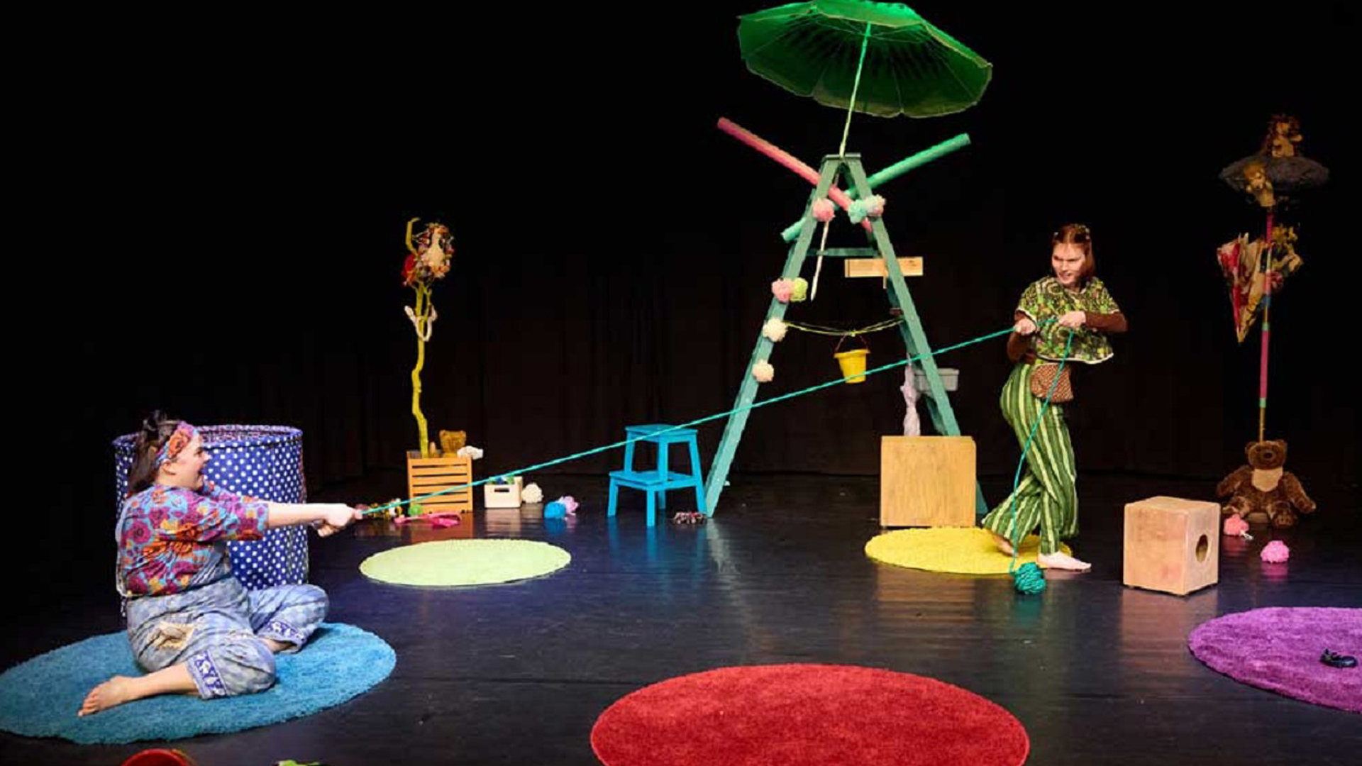Zwei Schauspielerinnen stehen auf einer farbenfrohen Theaterbühne. Sie halten ein grünes Seil, das sie spielerisch gegeneinander ziehen. Die Bühne ist mit bunten Teppichen, Requisiten aus Alltagsgegenständen und einer dekorierten Holzleiter gestaltet. Im Hintergrund ist ein schwarzer Vorhang zu sehen.