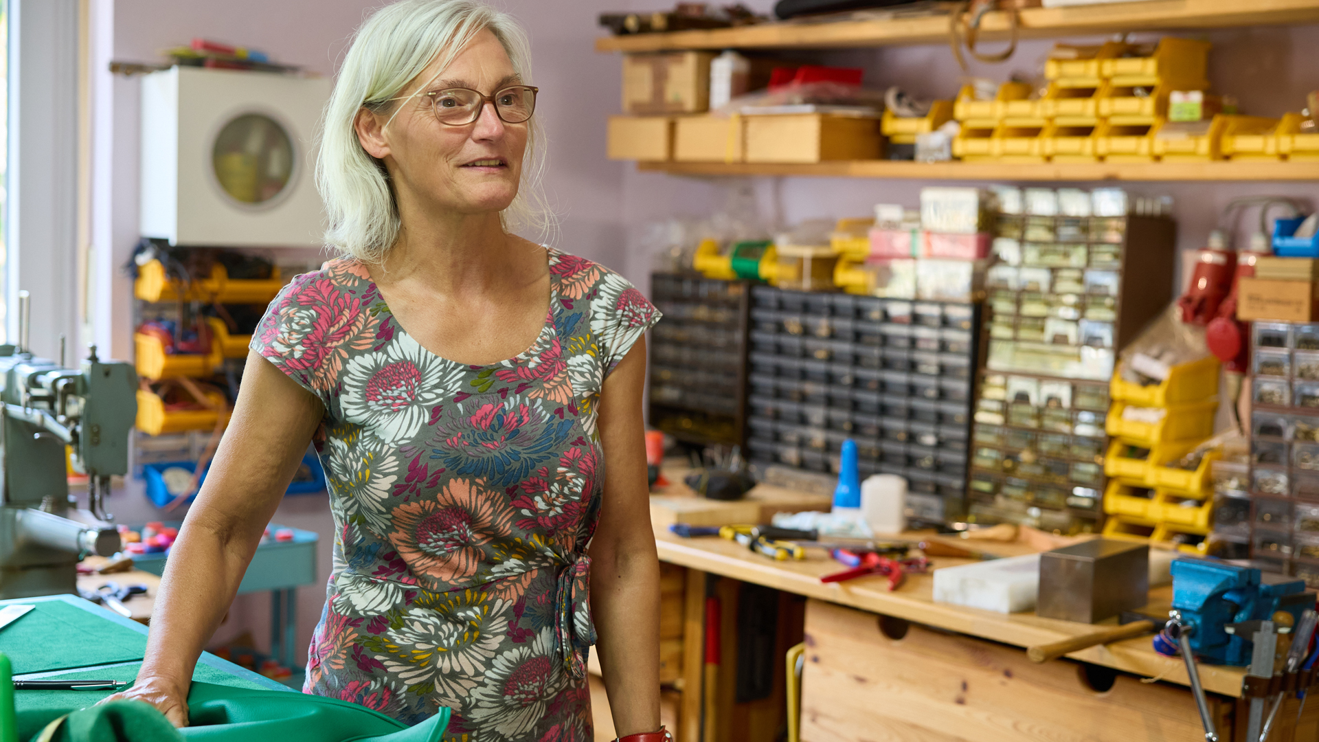 Ina May steht in ihrer Lederwerkstatt an der Luxemburger Straße in Sülz. Im Vordergrund liegt eine grüne Ledertasche auf dem Arbeitstisch. Im Hintergrund sind Regale mit zahlreichen Werkzeugen, Materialien und Kleinteilen zu sehen. Die Sattlerin trägt ein geblümtes Kleid und blickt lächelnd zur Seite.