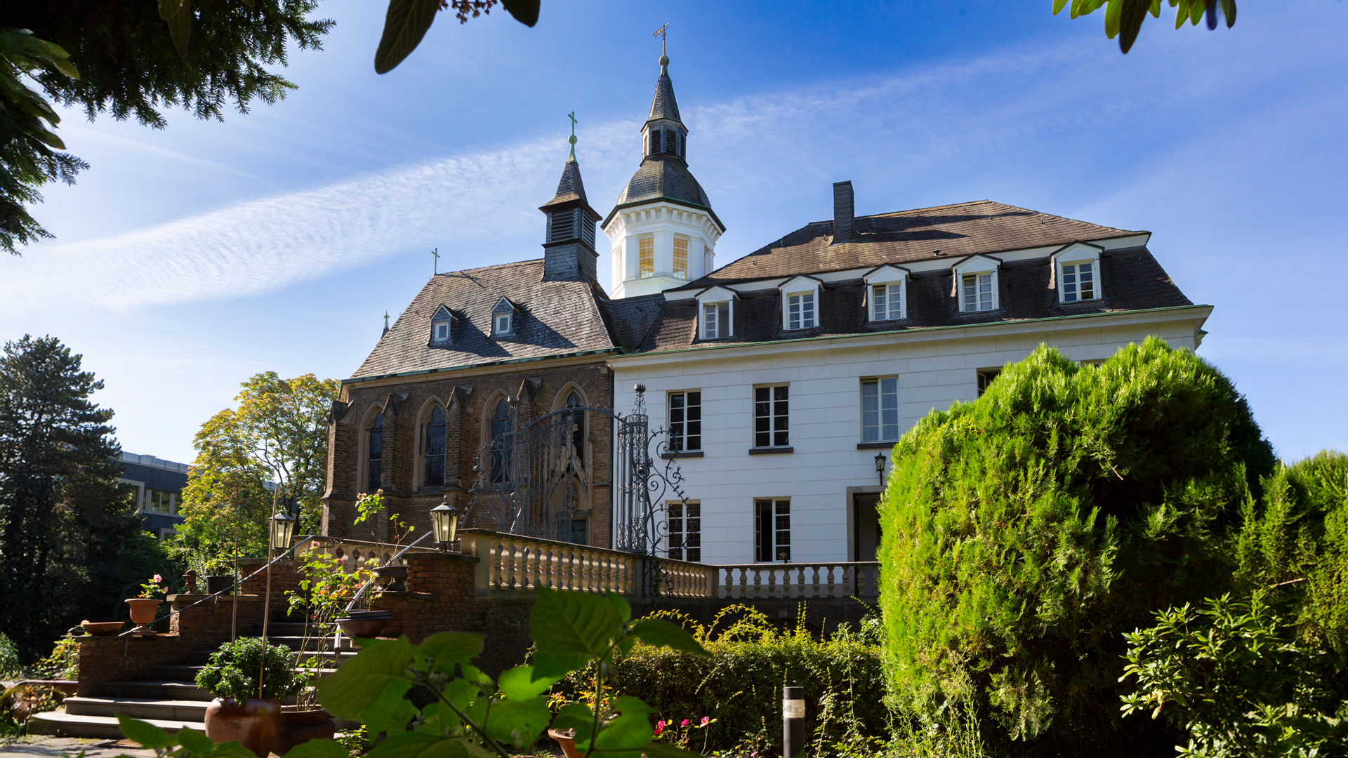 Das Weißhaus-Schlösschen in Köln bei strahlendem Sonnenschein, im Vordergrund üppiges Grün, im Hintergrund das historische Gebäude mit klassizistischem Anbau und Kapelle.